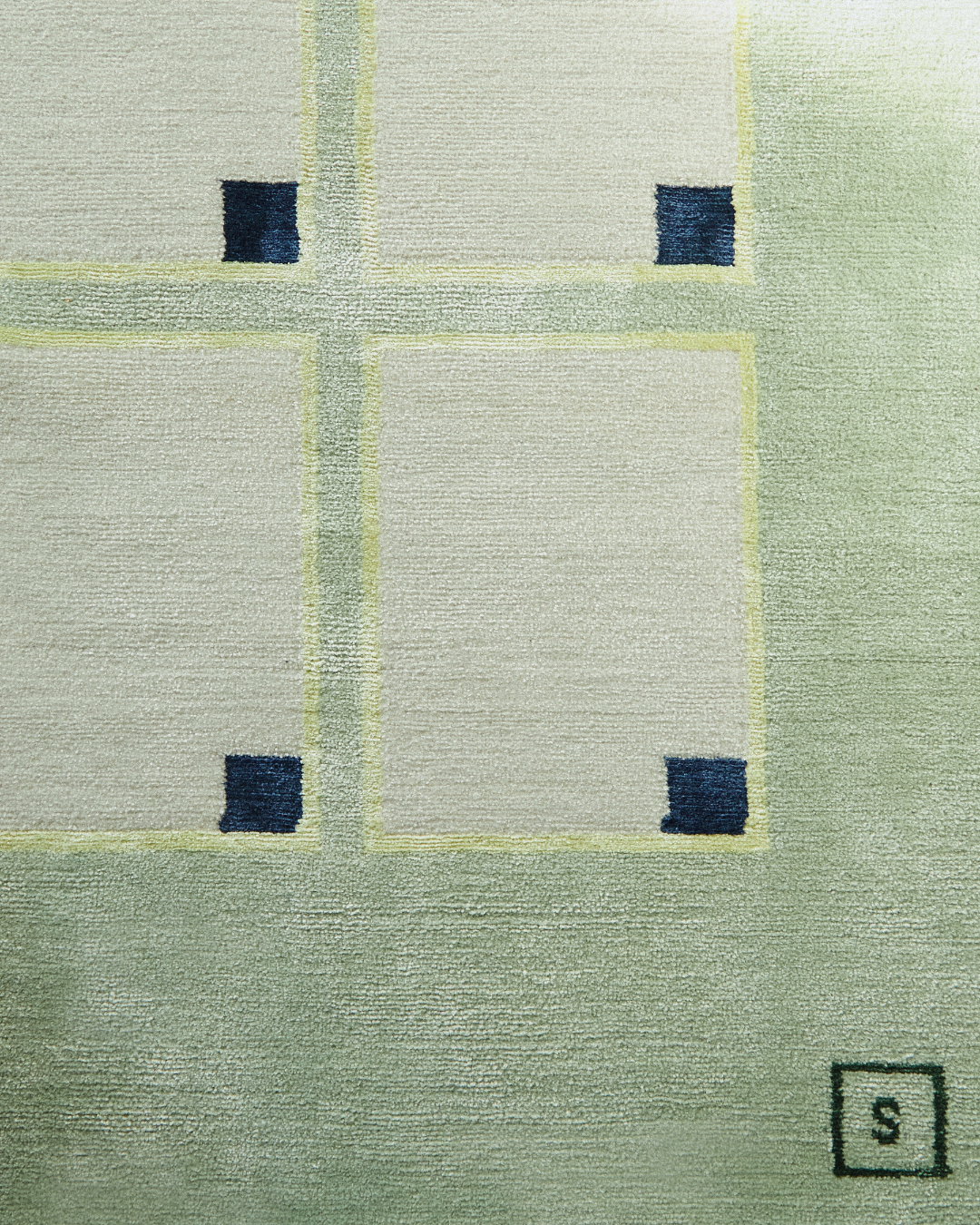 Detail image showcasing the geometric rectangular patterns on the pale green hand-knotted rug, featuring our signature 'S' logo in rich dark green.